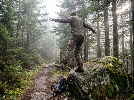Person balanciert auf einem Bein auf einem moosigen Felsen im Nebelwald mit Wanderschuhen – Gleichgewichtsübung zum Stärken der Knöchel beim Wandern