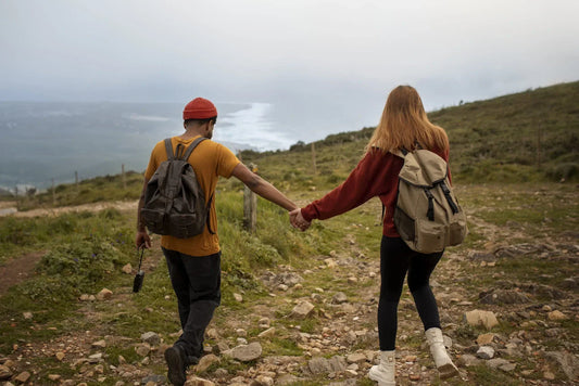 Paar von hinten beim Wandern auf einem felsigen Küstenpfad – Mann und Frau mit Rucksäcken Hand in Hand auf einem Trekking-Trail